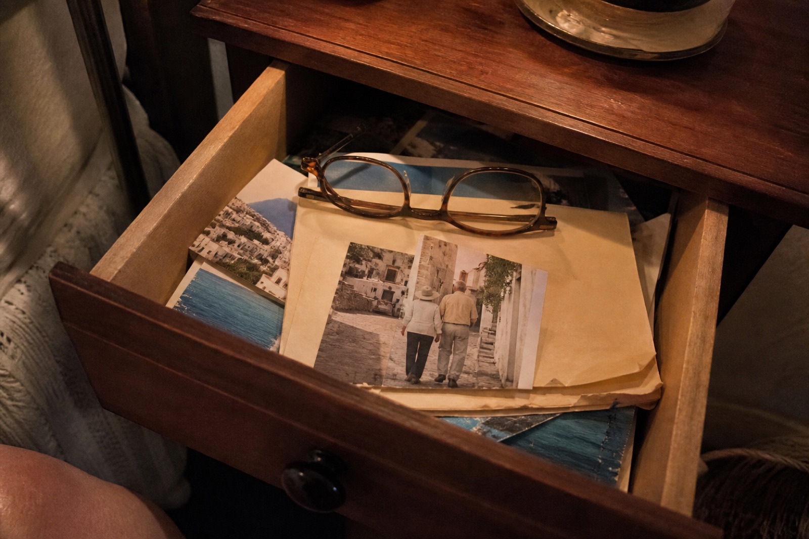 A nightstand drawer pulled open, showing a worn manila folder of Portugal clippings, screenshots, and an old cobblestone-street photo, with reading glasses on top